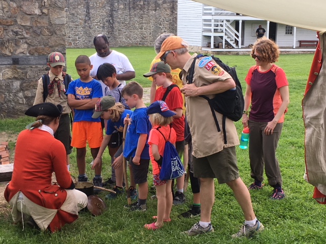 A group of kids looking over a solider who is cooking over a fire.