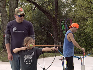 Man shows young girl how to hold a bow and arrow